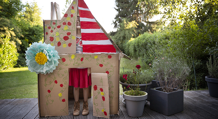 Child (6-7) playfully pulling a silly face through the gaps inside a homemade playhouse made from recycled cardboard